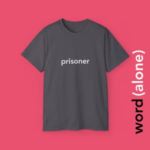 Könnte beinhalten: Ein dunkelgraues T-Shirt mit dem Wort "prisoner" in Weiß auf der Brust. Der Text "word (alone)" ist vertikal in Weiß auf der rechten Seite des Shirts aufgedruckt. Das Shirt wird vor einem rosa Hintergrund präsentiert.