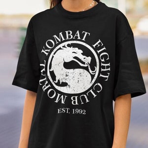 May include: Black t-shirt with a white graphic of the Mortal Kombat Fight Club logo. The logo features a dragon and the text "Mortal Kombat Fight Club" in a circle. The text "EST. 1992" is below the logo.