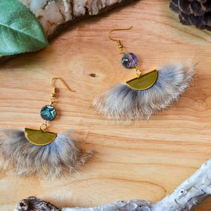May include: A pair of gold tone earrings with a feather design and iridescent abalone shell accents. The earrings are on a wooden surface.