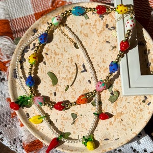 Peut inclure: Un collier coloré orné de perles en forme de fruits, notamment des fraises, des citrons et des piments. Le collier est composé de métal doré et de perles blanches. Le collier est présenté sur une assiette décorative.