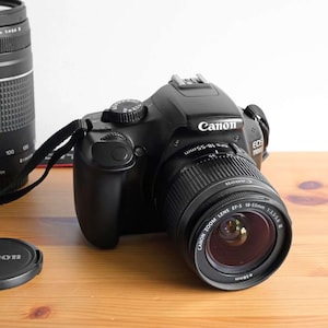 May include: A black Canon EOS 1100D digital SLR camera with an attached 18-55mm zoom lens, a separate 75-300mm telephoto lens, and a circular black Canon lens cap. The camera body features a mode dial, a hot shoe, and a black fabric neck strap.