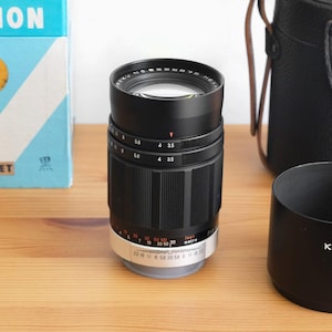 May include: A black Konica Hexanon Pre-Set lens with a silver ring and markings. The lens is 135mm and has an aperture of f/3.5. The lens is in a black leather lens case.