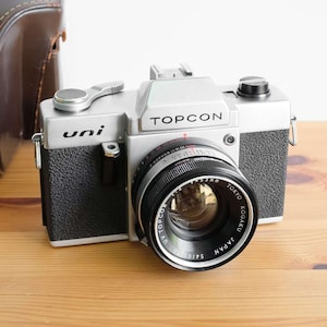 May include: A silver and black Topcon Uni camera with a lens from Tokyo, Japan, sits on a wooden surface. The camera has a black leather grip and a silver top with the brand name. A brown leather case is in the background.