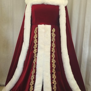 Puede incluir: Un disfraz de terciopelo rojo con ribete de piel sintética blanca y detalles bordados dorados. El disfraz incluye un gorro y una capa a juego, exhibidos en un maniquí. El atuendo está diseñado para una ocasión festiva.