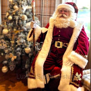 Op de afbeelding: Een kerstmankostuum van rood fluweel met witte bontkraag en gouden accenten. Het kostüm bestaat uit een lange jas, broek en een riem met een gouden gesp. De kerstman houdt een houten staf vast.