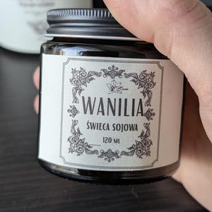 May include: A small amber glass jar with a black metal screw-top lid and a white rectangular label. The label features decorative black filigree borders and the text WANILIA, SWIECA SOJOWA, and 120 ML printed in a clear, dark font.