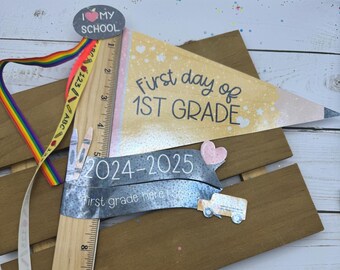 以下が含まれることがあります： 「First day of 1st Grade」と書かれた鉛筆の形をした旗と、「2024-2025 first grade here I come」と書かれたハートがついた木製の定規。虹色のリボンと、「I love my school」と書かれた赤いハートとリンゴのついた小さな丸いステッカーがあります。