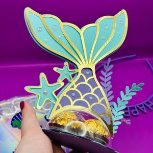 Könnte beinhalten: Eine türkisfarbene und goldene Meerjungfrauen-Schwanz-Geschenkbox mit einer durchsichtigen Plastikkuppel. Die Box ist mit goldenem Glitzer und weißen Perlen verziert. Die Box ist mit zwei Schokoladenbonbons gefüllt.