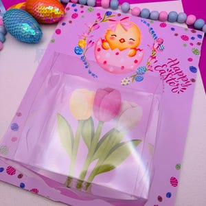 May include: An Easter-themed gift set featuring a clear plastic box containing three tulip-shaped decorations in yellow, pink, and white. The set is presented on a lavender card with a cartoon chick hatching from an egg and the words "Happy Easter". Decorative sequined eggs are also visible.