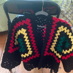 May include: A black crochet cardigan with a colorful pattern. The design features red, yellow, and green squares, creating a vibrant and eye-catching look. The cardigan is displayed on a hanger.