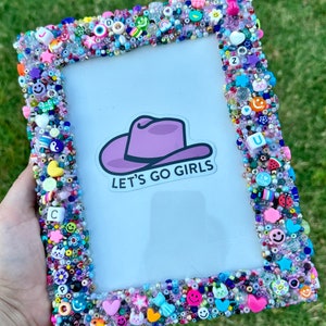 May include: A purple picture frame decorated with colorful beads, plastic charms, and letters. The frame has a white background with a sticker of a purple cowboy hat and the text "Let's Go Girls".