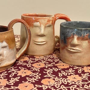 May include: Three handmade ceramic mugs featuring sculpted human faces on the front. The left mug has a tan and white glaze, the center mug is a light peach tone, and the right mug features a blue and tan glaze. Each mug has a curved handle.