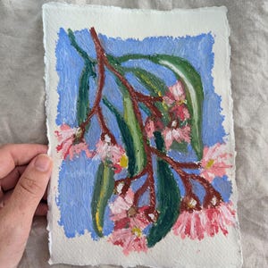 以下が含まれることがあります： ピンクの花と緑の葉を持つ、青い背景の開花した枝のオリジナルペインティング。アートワークは、デッケルエッジのあるテクスチャード加工された白い紙に描かれています。絵は手で持たれています。