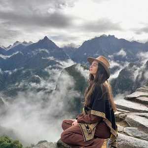 May include: A person wearing a brown hat, black poncho with gold geometric patterns, and brown trousers sits on a rock with eyes closed. The background features a mountain range with clouds and fog.