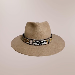 May include: A tan fedora-style hat with a decorative band. The band features a pattern of black, white, and gold beads. The hat has a wide brim and a rounded crown. The hat is a neutral color and would be suitable for many outfits.