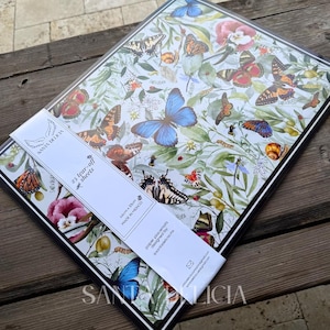 May include: A rectangular pad of 25 tear-off paper placemats featuring a botanical print with various colourful butterflies, green leaves, and flowers. A white paper band across the centre displays the text Santa Delicia, 25 tear-off sheets, 44cm x 33cm.