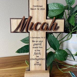 May include: Wooden cross with the name "Micah" in a cursive font. The cross has a light wood center with a darker wood border. Text includes "Confirmed March 12, 2024" and a Bible verse from 1 Corinthians 16:13.