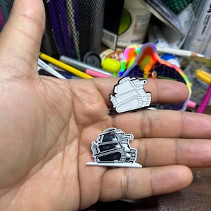Peut inclure: Deux épingles en émail représentant un char de bande dessinée, l'une noire et l'autre blanche. Les épingles sont tenues dans une main.