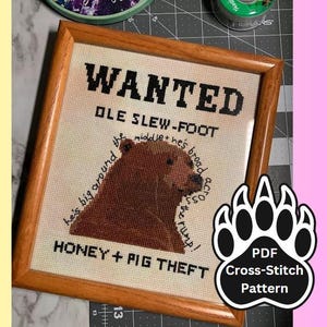 May include: A cross-stitch pattern for a wanted poster featuring a cartoon bear with the text "WANTED OLE SLEW-FOOT he's big around the middle + he's broad across the rump! HONEY + PIG THEFT". The pattern is in a wooden frame and a black paw print with the text "PDF Cross-Stitch Pattern" is in the lower right corner.
