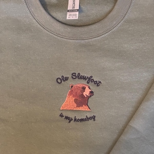 May include: A green sweatshirt with an embroidered brown bear and the text "Ole Slewfoot is my homeboy".