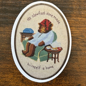 Può includere: Una toppa rotonda ricamata con un orso marrone che indossa un cappello rosso e una veste blu seduto su una sedia. Il testo intorno all'orso dice "ole slewfoot done made himself a home".