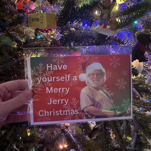 May include: A Christmas card with the text "Have yourself a Merry Jerry Christmas" featuring a man in a Santa hat playing a guitar. The card is held in front of a decorated Christmas tree.