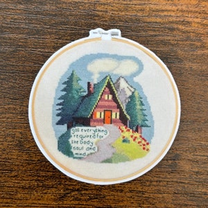 以下が含まれることがあります： 白い背景に、森の中の小屋がカラフルに描かれたクロスステッチの刺繍枠。小屋には赤いドアと煙突があり、煙が出ています。背景には木々と山があります。小屋の下には「got everything required for the body soul and mind」という文字が刺繍されています。