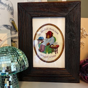 Puede incluir: Un bordado de punto de cruz enmarcado con un oso con un sombrero rojo sentado en una silla con una taza de té. El texto dice "Ole Slewfoot done made himself a home".