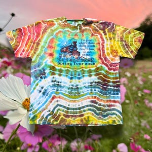 Puede incluir: Una camiseta teñida con un diseño de tren y el texto "I'm goin' where these chilly winds don't blow, oh me, oh my... Spring Tour 2026." La camiseta presenta una vibrante mezcla de rojo, amarillo, azul y blanco.