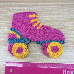 May include: A punch needle roller skate patch featuring a bright pink boot with yellow laces, teal accents, and yellow wheels with pink centers. The skate is positioned above a pink ruler showing a width of approximately 5.5 inches.