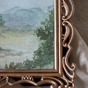 May include: A watercolor painting of a landscape with mountains and trees, framed in a gold ornate frame.