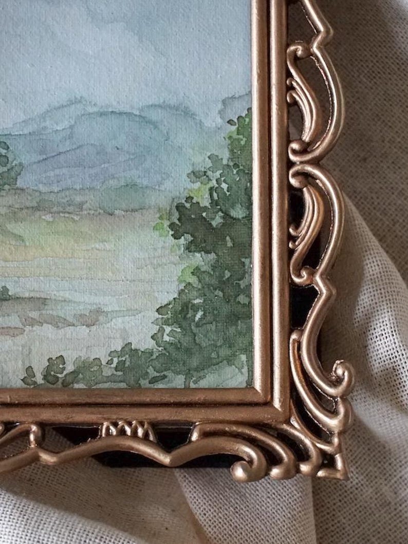 May include: A watercolor painting of a landscape with mountains and trees, framed in a gold ornate frame.