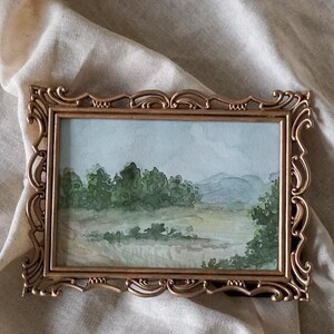 May include: A framed watercolor painting of a landscape with trees and hills in the background. The frame is gold and has a decorative design.