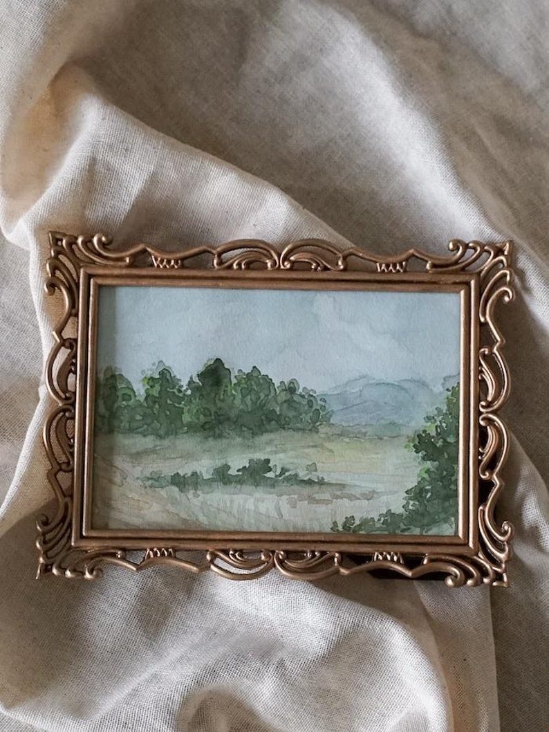 May include: A framed watercolor painting of a landscape with trees and hills in the background. The frame is gold and has a decorative design.