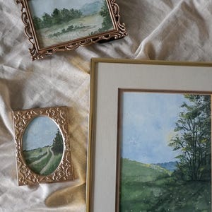 May include: Three framed watercolor paintings of landscapes. The paintings are in gold frames with ornate details. The paintings depict a winding road, a forest, and a field of wildflowers.