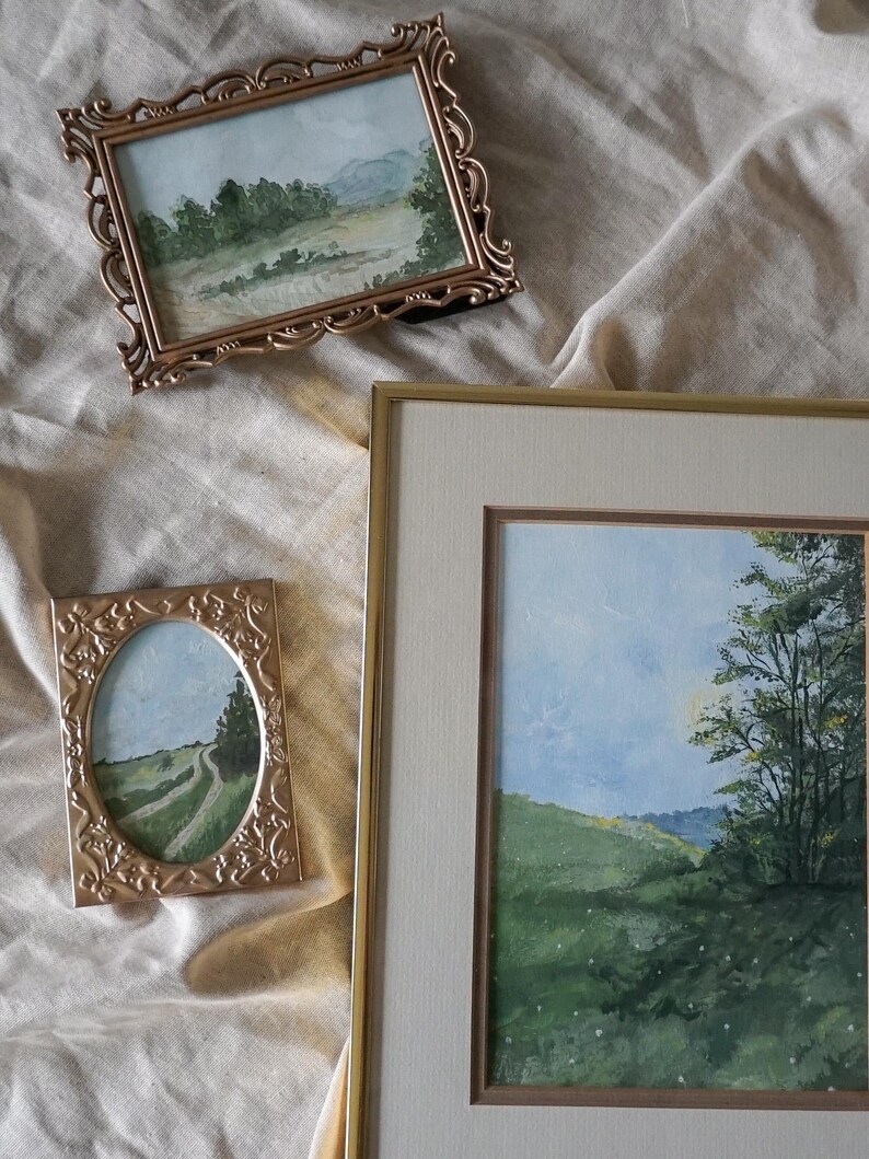 May include: Three framed watercolor paintings of landscapes. The paintings are in gold frames with ornate details. The paintings depict a winding road, a forest, and a field of wildflowers.