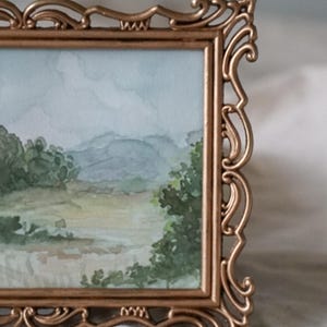 May include: A gold-toned ornate frame with a watercolor painting of a landscape scene. The painting depicts a field with trees and mountains in the background.