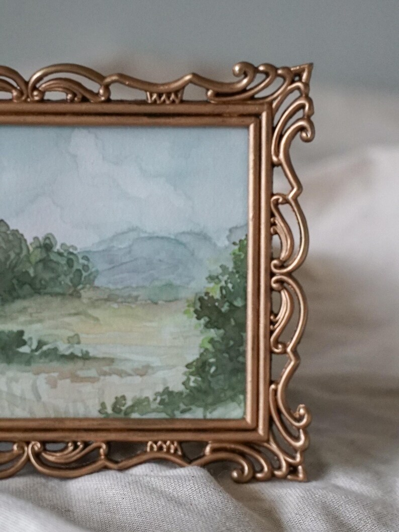 May include: A gold-toned ornate frame with a watercolor painting of a landscape scene. The painting depicts a field with trees and mountains in the background.