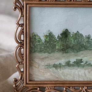 May include: A watercolor painting of a landscape scene framed in a gold ornate frame. The painting depicts a field of green trees and a light blue sky.