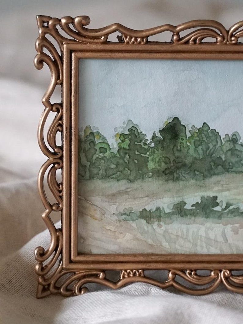 May include: A watercolor painting of a landscape scene framed in a gold ornate frame. The painting depicts a field of green trees and a light blue sky.