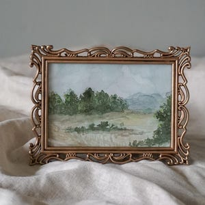 May include: A watercolor painting of a landscape scene with trees, hills, and a field, framed in a gold ornate frame.