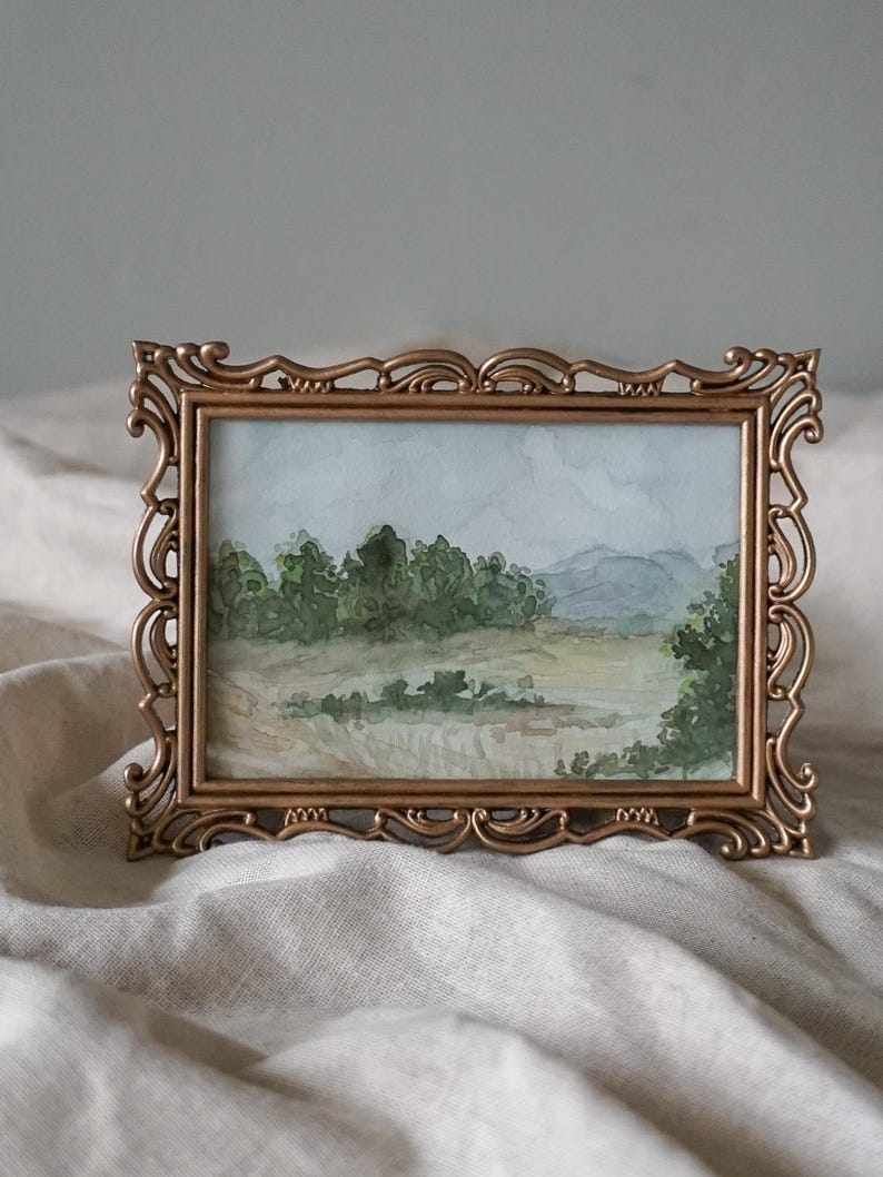 May include: A watercolor painting of a landscape scene with trees, hills, and a field, framed in a gold ornate frame.