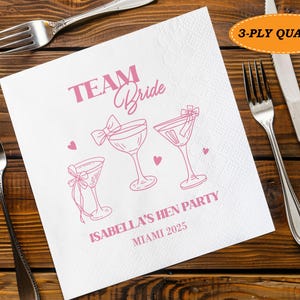 May include: White paper napkin with pink text and illustrations. The text reads "TEAM Bride" and "ISABELLA'S HEN PARTY MIAMI 2025". The napkin features drawings of cocktail glasses and hearts. The napkin is 3-ply quality.