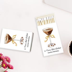 May include: Two white matchboxes with a watercolor illustration of a cocktail, a yellow bow, and coffee beans. One box is open, revealing the matches. Text reads "a Tini Bit Older Amelia's Birthday".