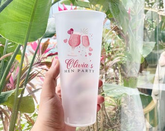 May include: A tall, frosted plastic cup with a pink ombre effect. The cup is decorated with a cocktail and champagne glass illustration, hearts, and the text "Olivia's HEN PARTY" in pink script. The cup is held by a person.