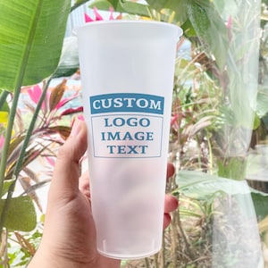 Puede incluir: Un vaso alto de plástico esmerilado con una etiqueta rectangular azul y blanca que dice "CUSTOM LOGO IMAGE TEXT". El vaso se sostiene frente a un fondo de plantas verdes y rosas.
