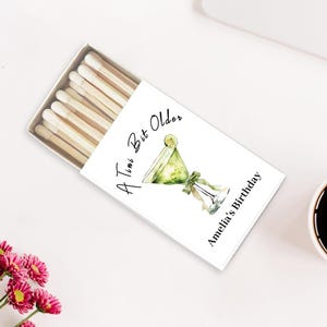 May include: A white matchbox with the text "A Time But Older" and "Amelia's Birthday" next to a watercolor illustration of a green cocktail. The matchbox is open, revealing several wooden matches with white tips. A cup of coffee and pink flowers are also in the image.