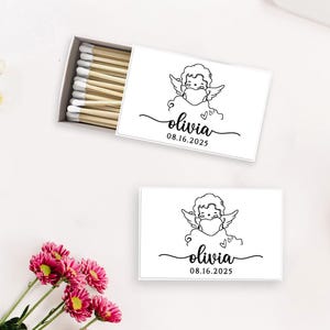 May include: White matchboxes with a black line drawing of an angel wearing a mask, the name "olivia" in cursive, and the date "08.16.2025". One box is open, revealing the matches. Pink and white flowers are in the background.