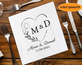 Può includere: Tovagliolo di carta bianco con un motivo floreale a forma di cuore, le iniziali "M & D" e i nomi "Maria & Daniel" con la data "11.09.2025". Posate circondano il tovagliolo su un tavolo di legno. Il testo "3-PLY QUALITY" è in un ovale arancione.
