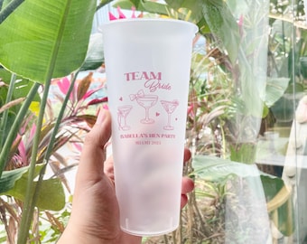 May include: A frosted plastic cup with pink text and illustrations. The text reads "Team Bride" above three cocktail glasses and "Isabella's Hen Party Miami 2025". The cup is held in front of a background of green leaves and pink flowers.